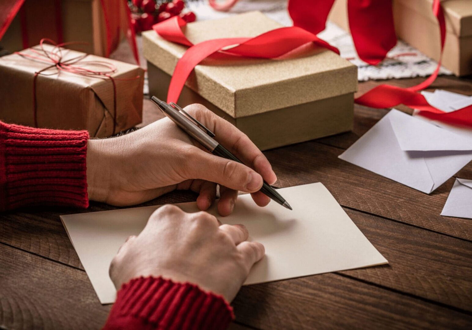 Hands writing holiday card beside wrapped gifts